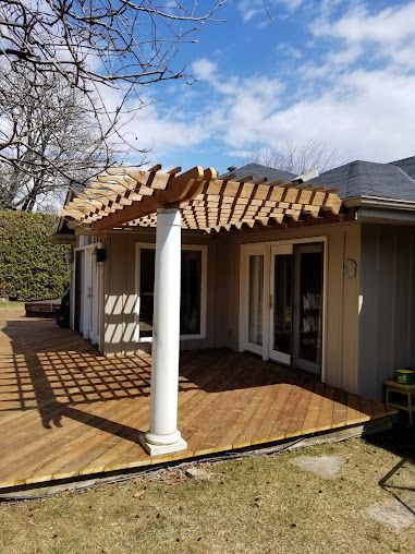 A pergola is sitting on top of a wooden deck in front of a house.