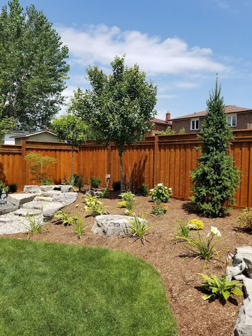 A wooden fence surrounds a lush green yard with trees and rocks.