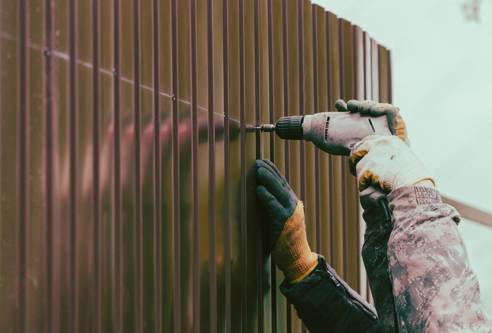 A worker in work gloves uses a power drill to fasten a brown corrugated metal panel to a wall.