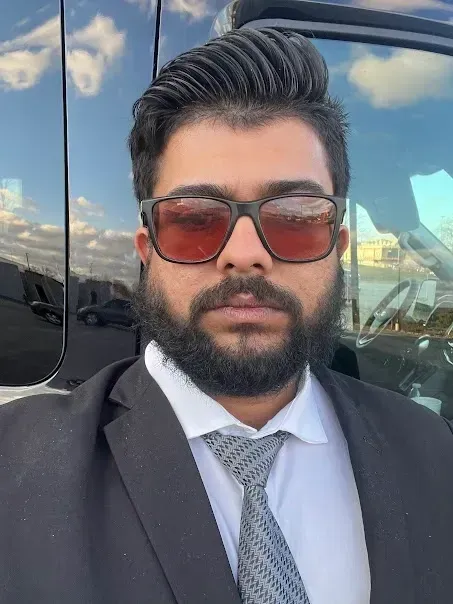 A man with a beard wearing sunglasses and a suit is standing in front of a car.