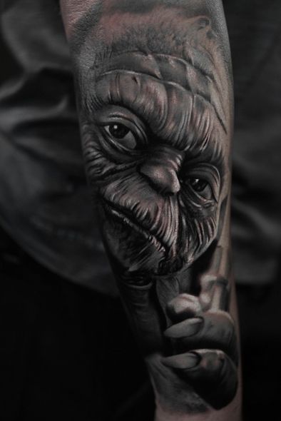 Realistic black and grey tattoo of Yoda from Star Wars on a forearm. Shows Yoda's face and hand holding a lightsaber.