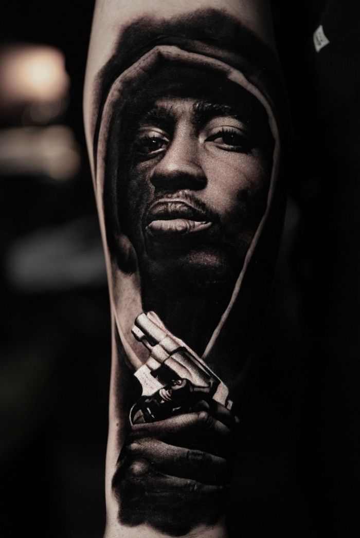 Black and white tattoo of Tupac holding a gun, in a hooded sweatshirt.