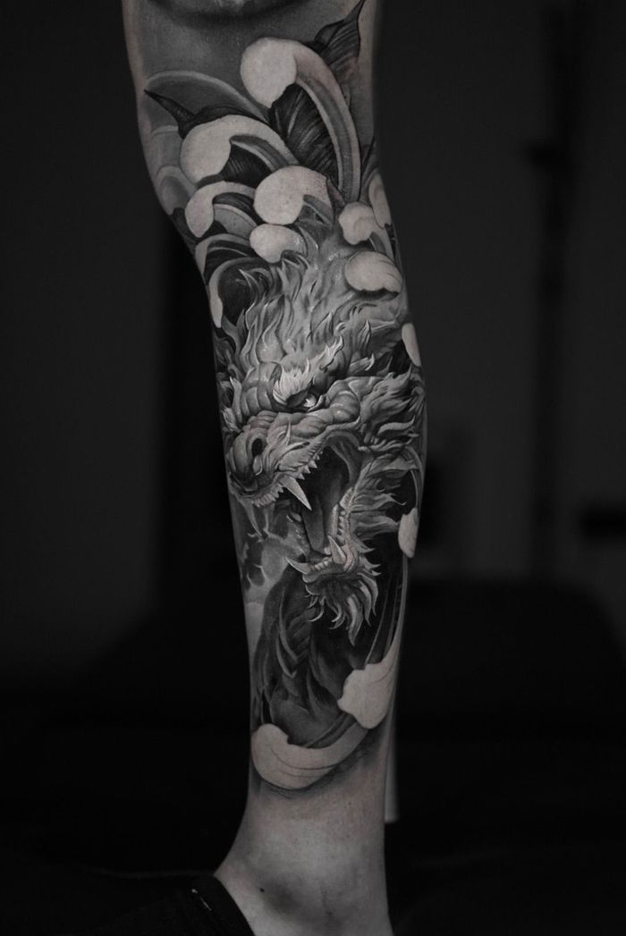 Grayscale tattoo on a leg — fierce dragon's head with open mouth surrounded by stylized water waves.