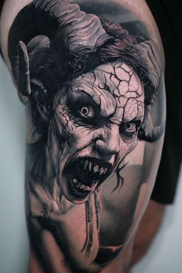Detailed black and gray tattoo of a monstrous, horned face with an open mouth, exposed teeth, and cracked skin.