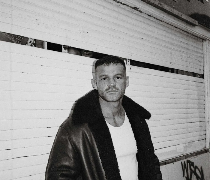 Joe  in coat, white tank top, stands in front of a white shuttered wall, looking directly at the camera.