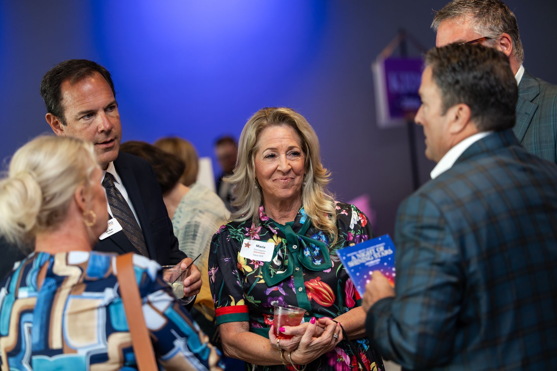 People at a social gathering, engaged in conversation. Woman in floral dress smiling, holding drink.