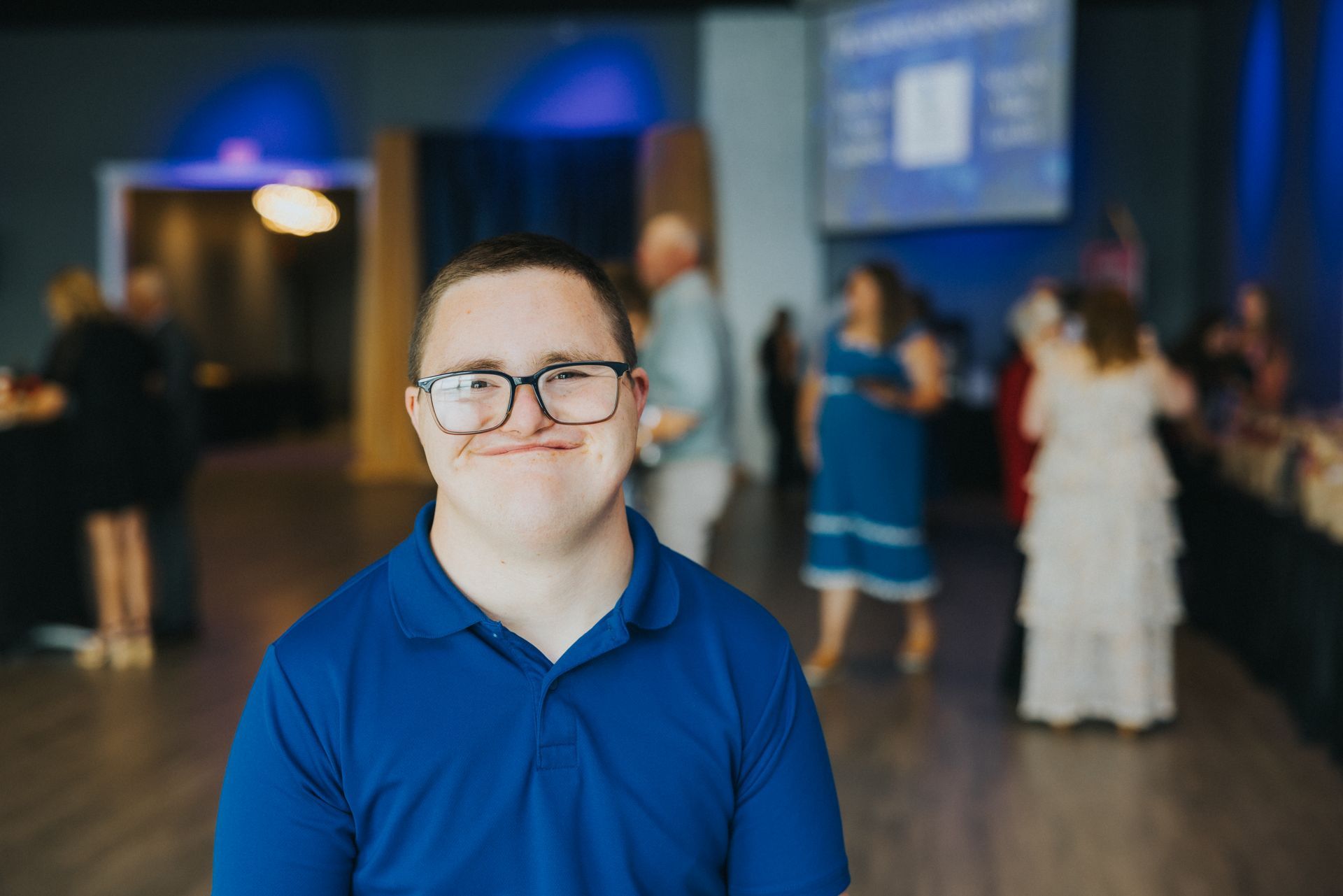 A smiling person with glasses in a blue shirt at an event with people in the background.