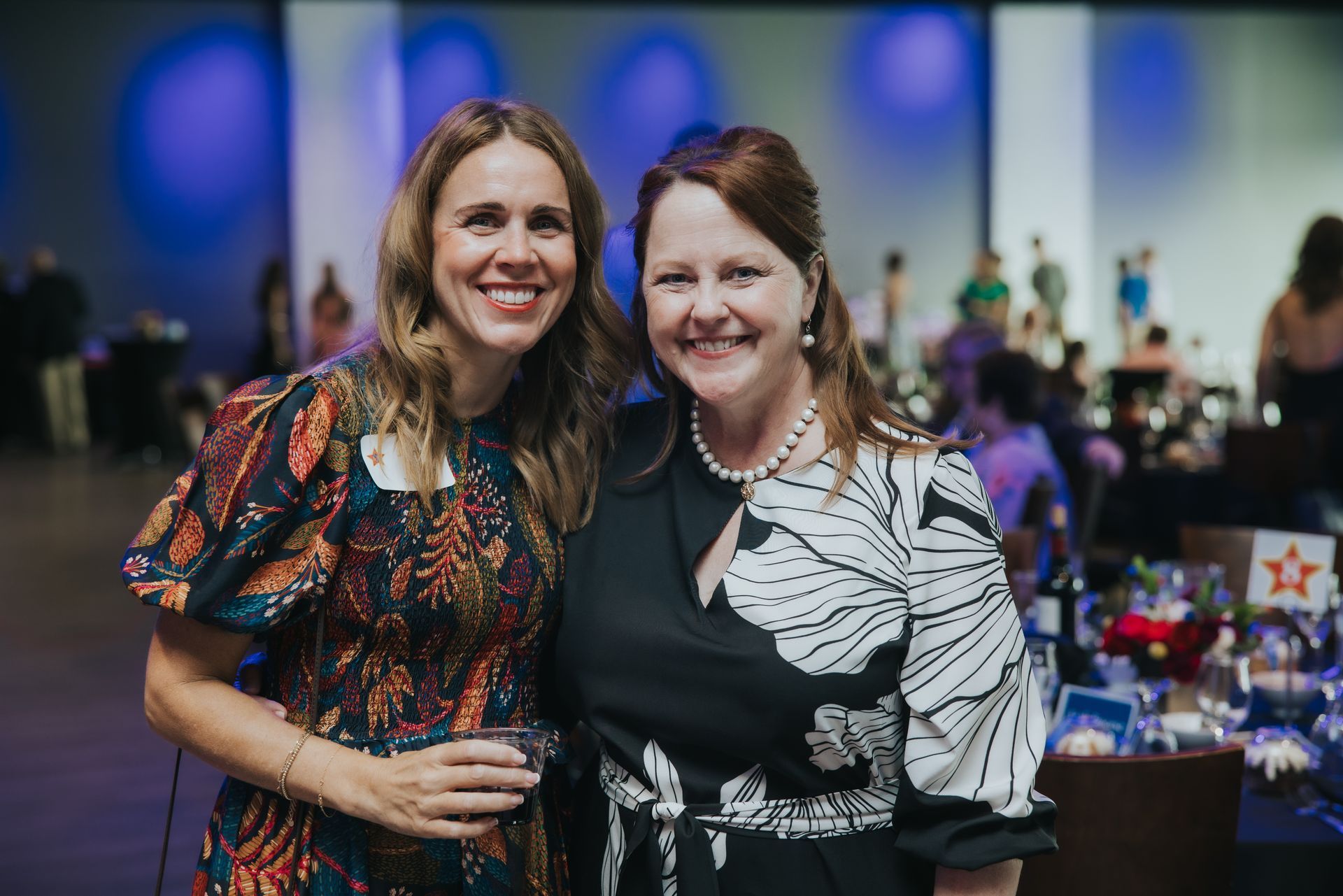 Two women smiling at a formal event; one in a patterned dress, the other in a black and white top.