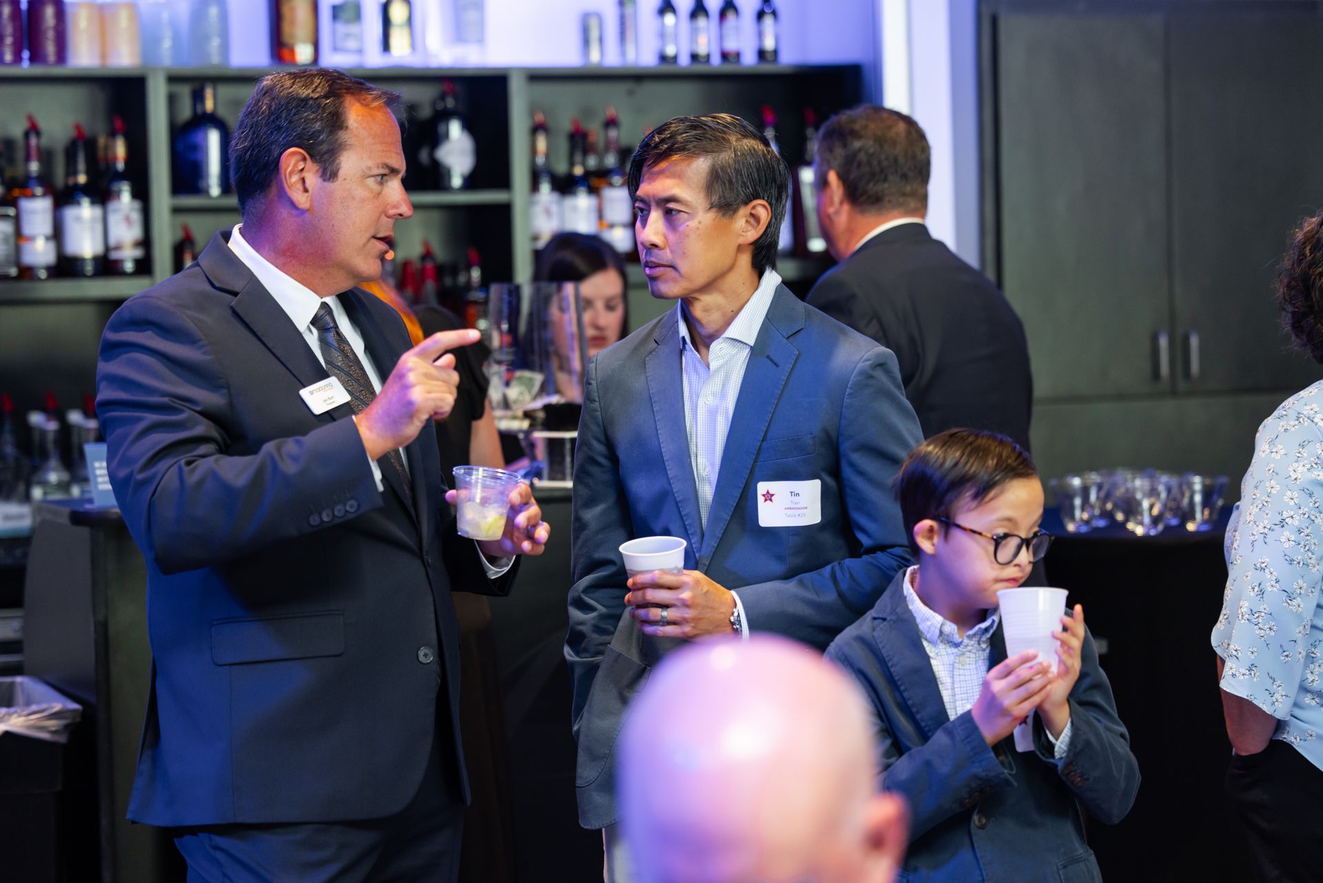 Men in suits and a young person in a suit converse and drink at an event; bar in background.