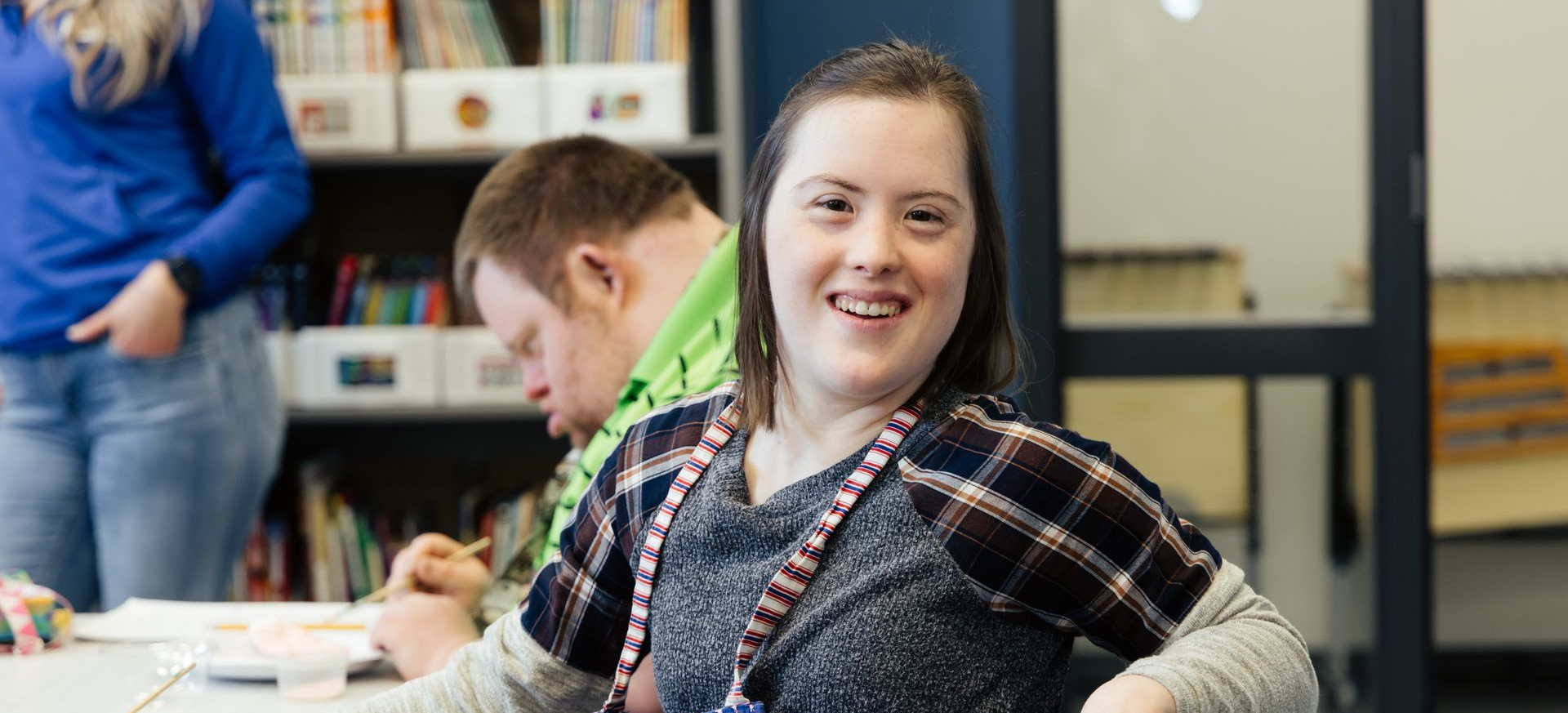 A smiling woman with Down syndrome sits in a classroom. A man with Down syndrome is nearby.