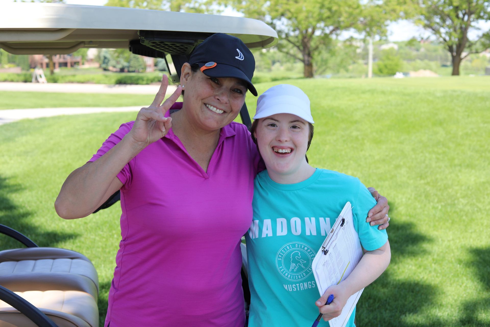 Two women smiling, posing for photo on golf course; one gives peace sign.