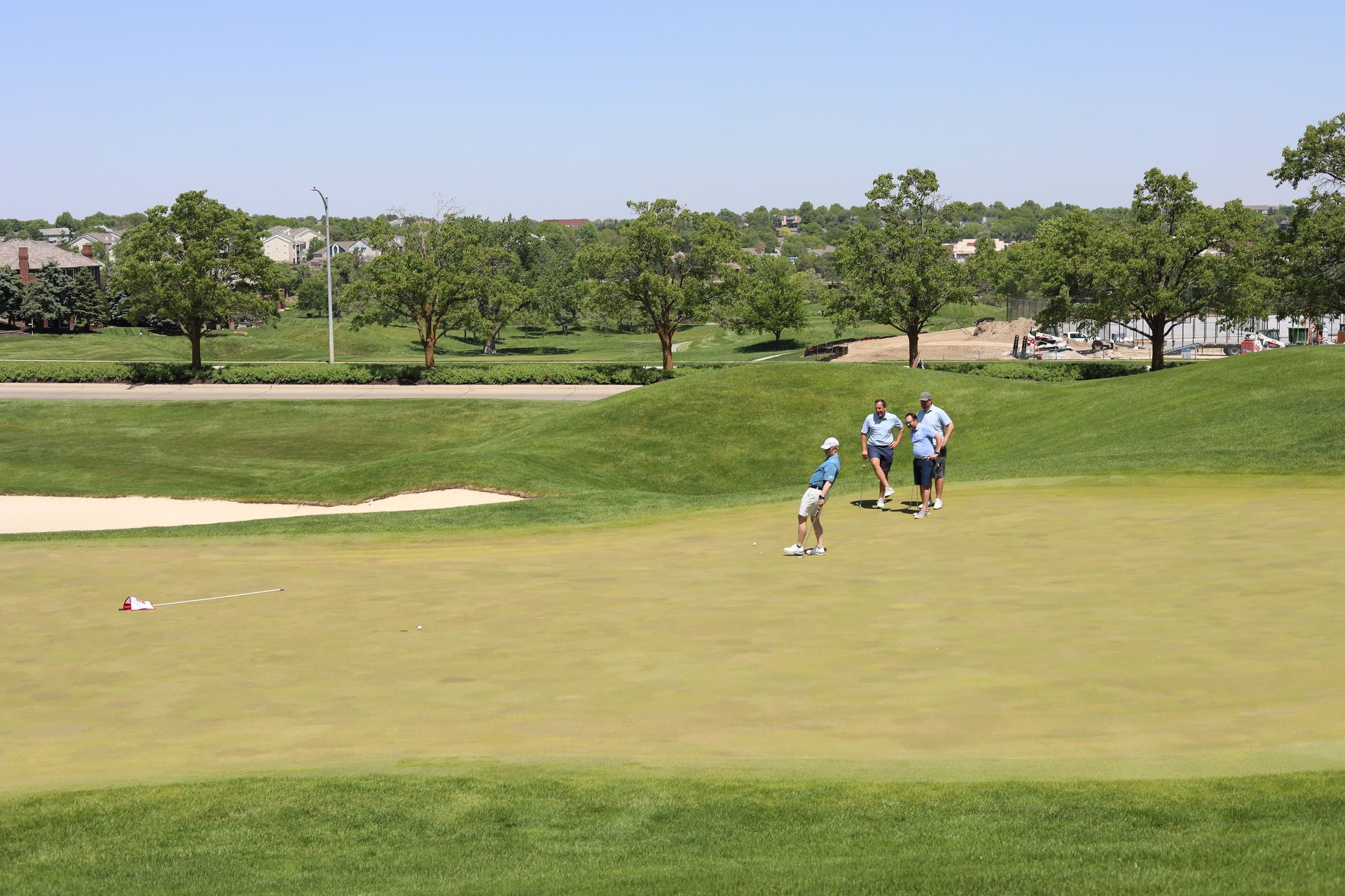 People playing golf on a green course under a blue sky, with trees in the background.
