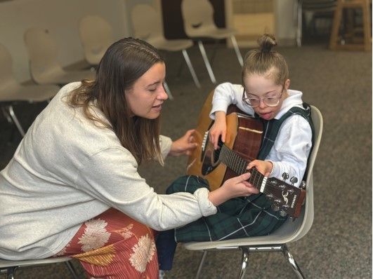 Woman teaching a person with Down syndrome to play guitar. They are in a room with chairs.
