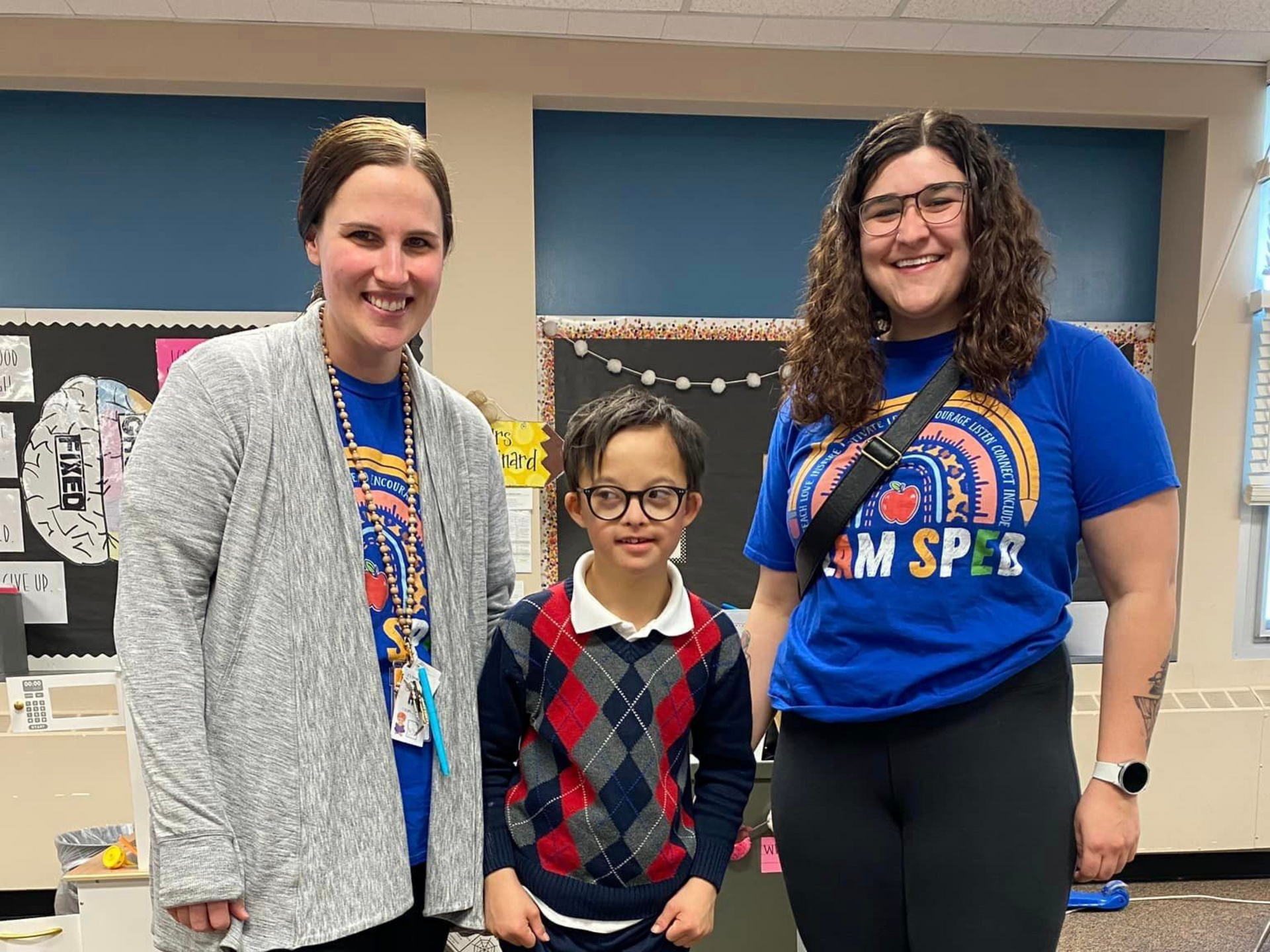 Two adults flank a child in a classroom. All three smile. The child wears glasses and a sweater vest.