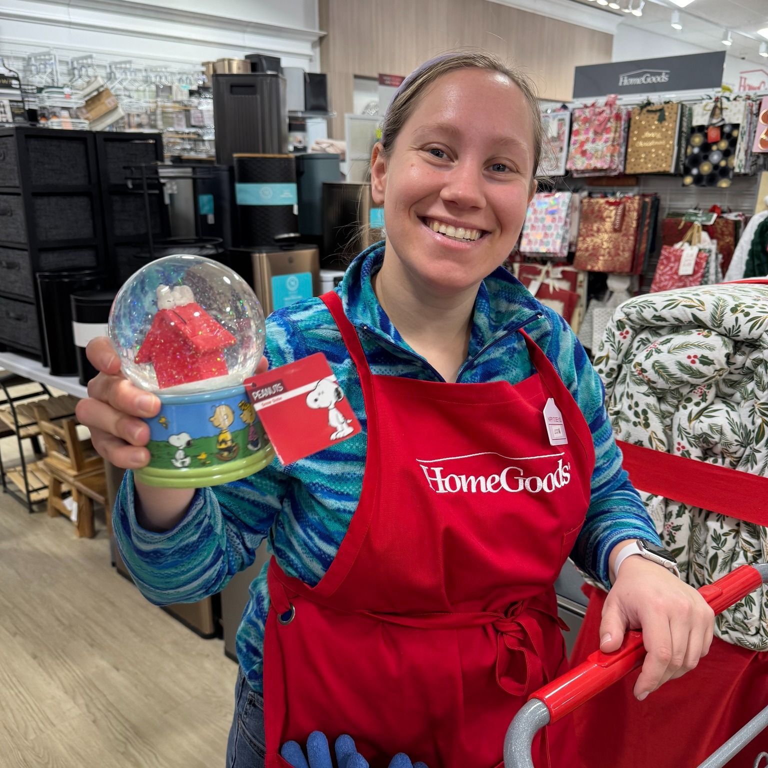 Woman in red apron at HomeGoods holding a Snoopy snow globe.