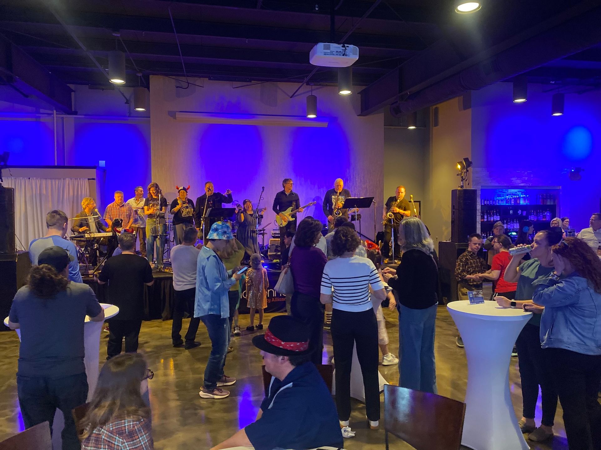 Band performing on stage in a venue with an audience, blue lighting, and small tables.