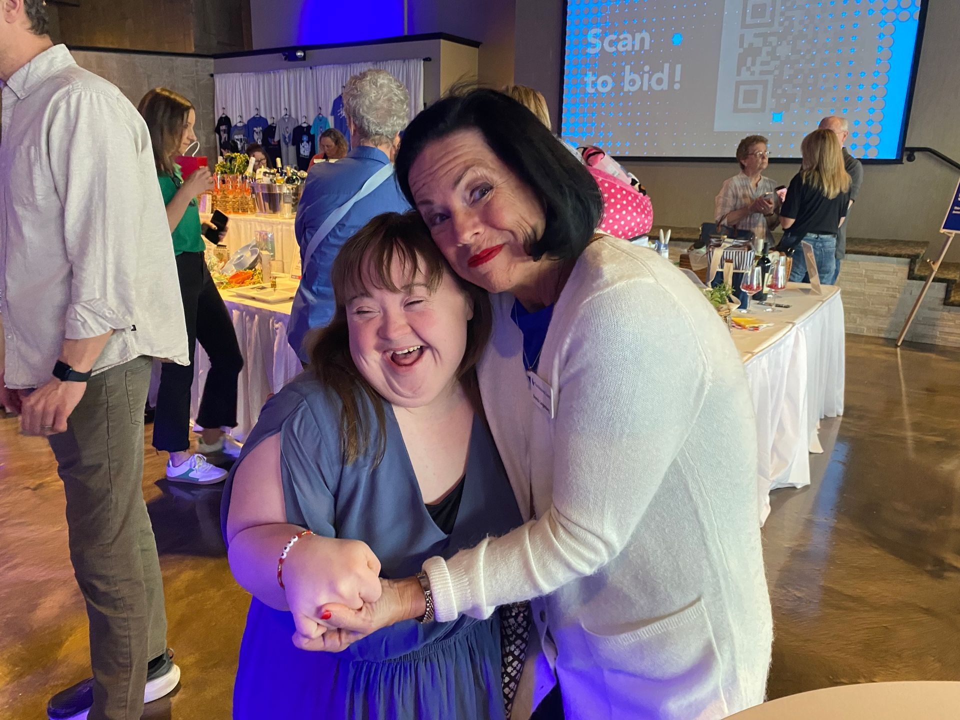 Two women embracing, smiling. One with Down syndrome, in a blue dress. They're at an event, near tables.
