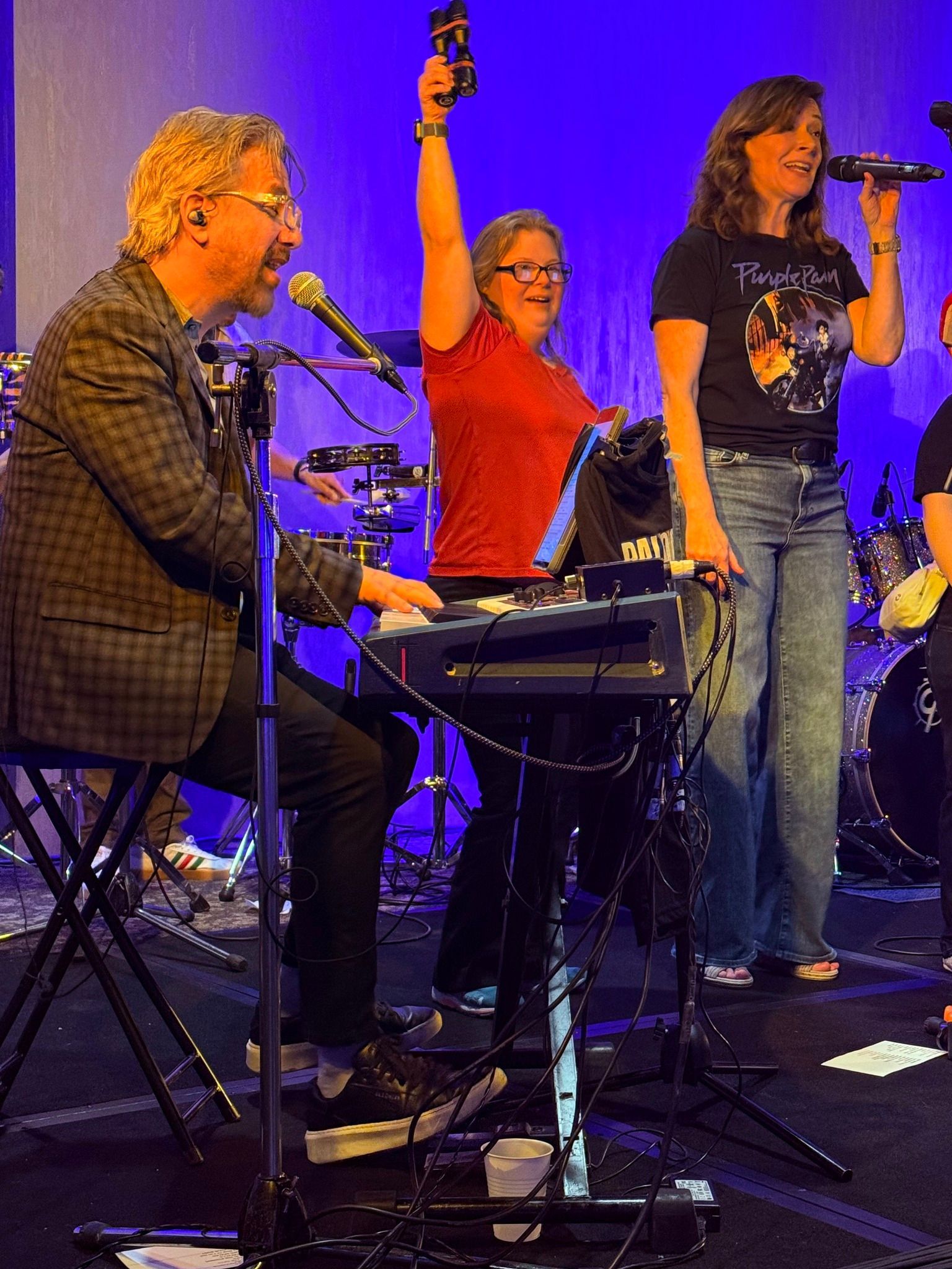 A band performing on stage. A man plays keyboard, a woman sings with a microphone, and another holds up a tambourine.