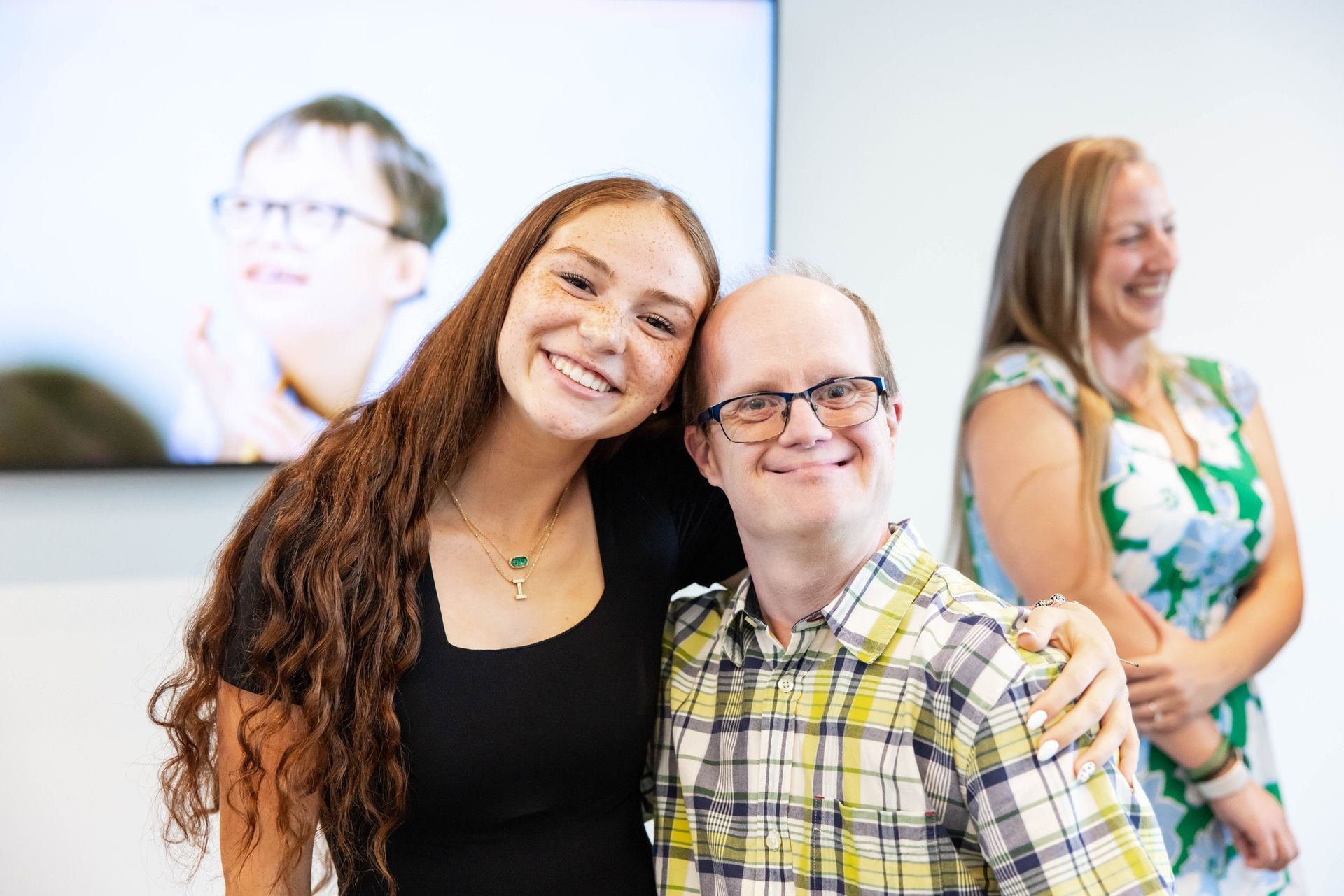 Woman with long hair hugs a man with Down syndrome, both smiling. A third woman smiles in the background.
