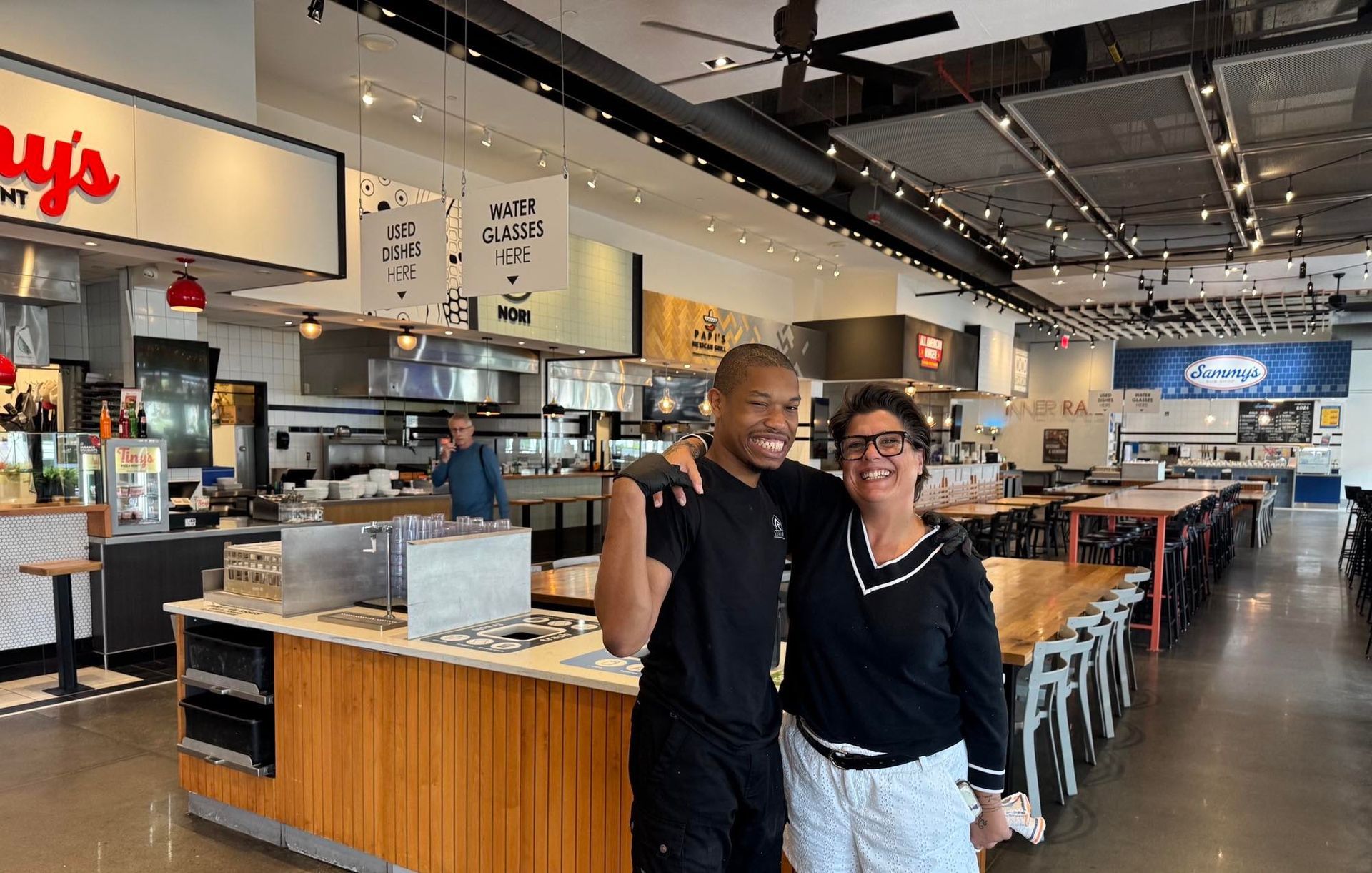 Two people smiling with arms around each other inside a restaurant. Open kitchen and tables in the background.