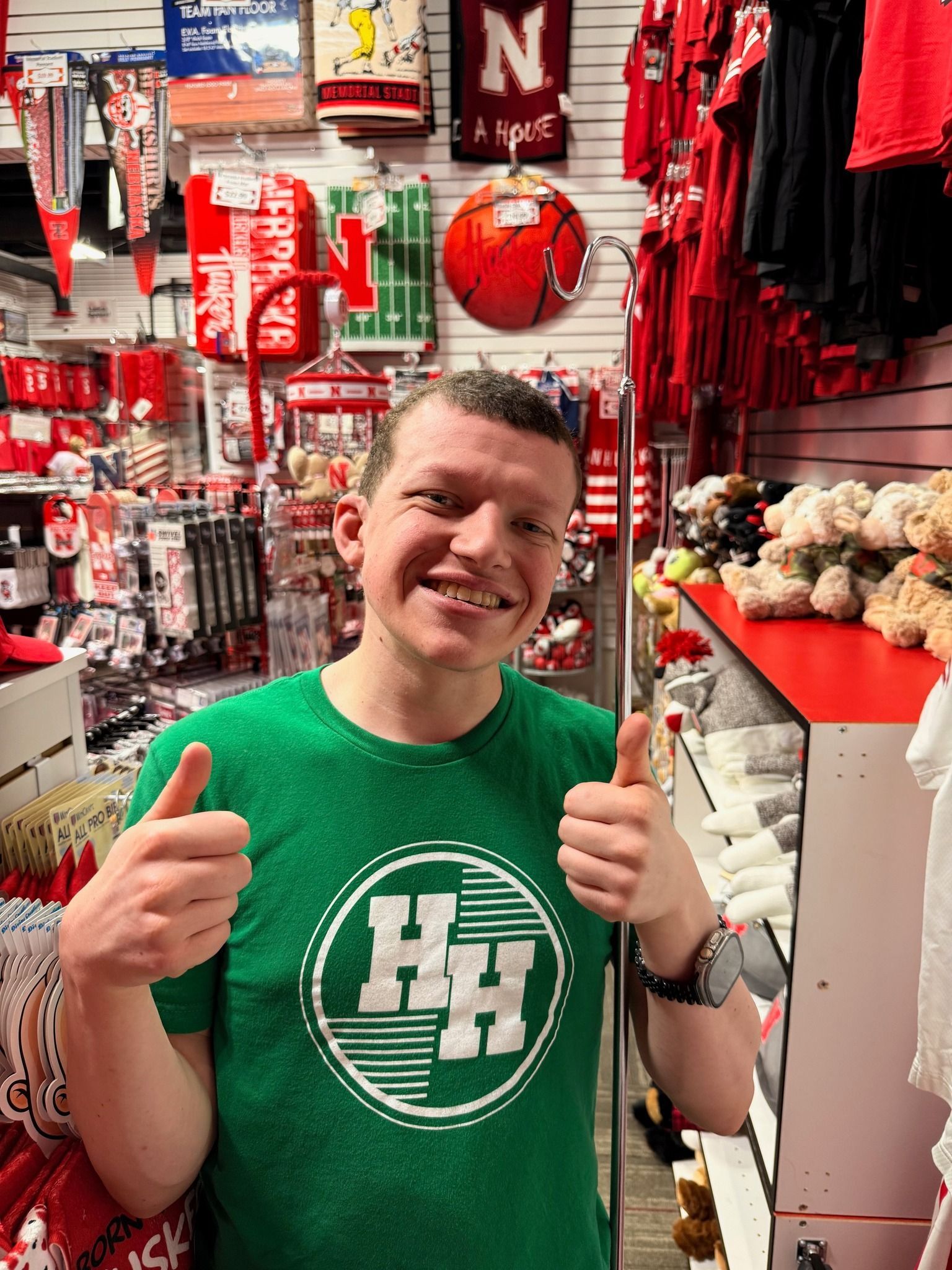 Man in green shirt gives thumbs up in a Nebraska Huskers shop. Red and white merchandise surrounds.