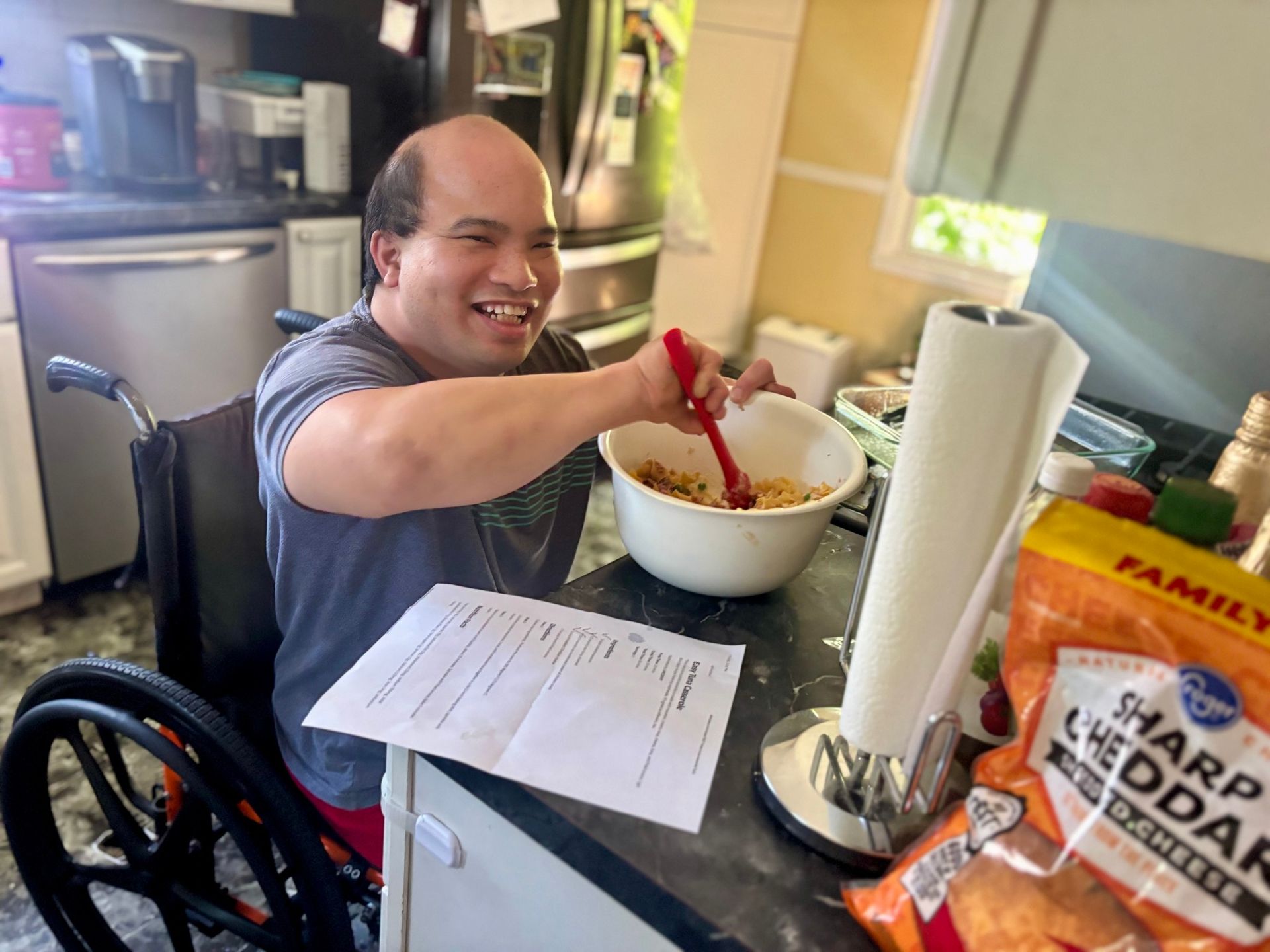 Man in wheelchair mixing in a bowl, smiling. Kitchen setting, bag of cheddar cheese visible.