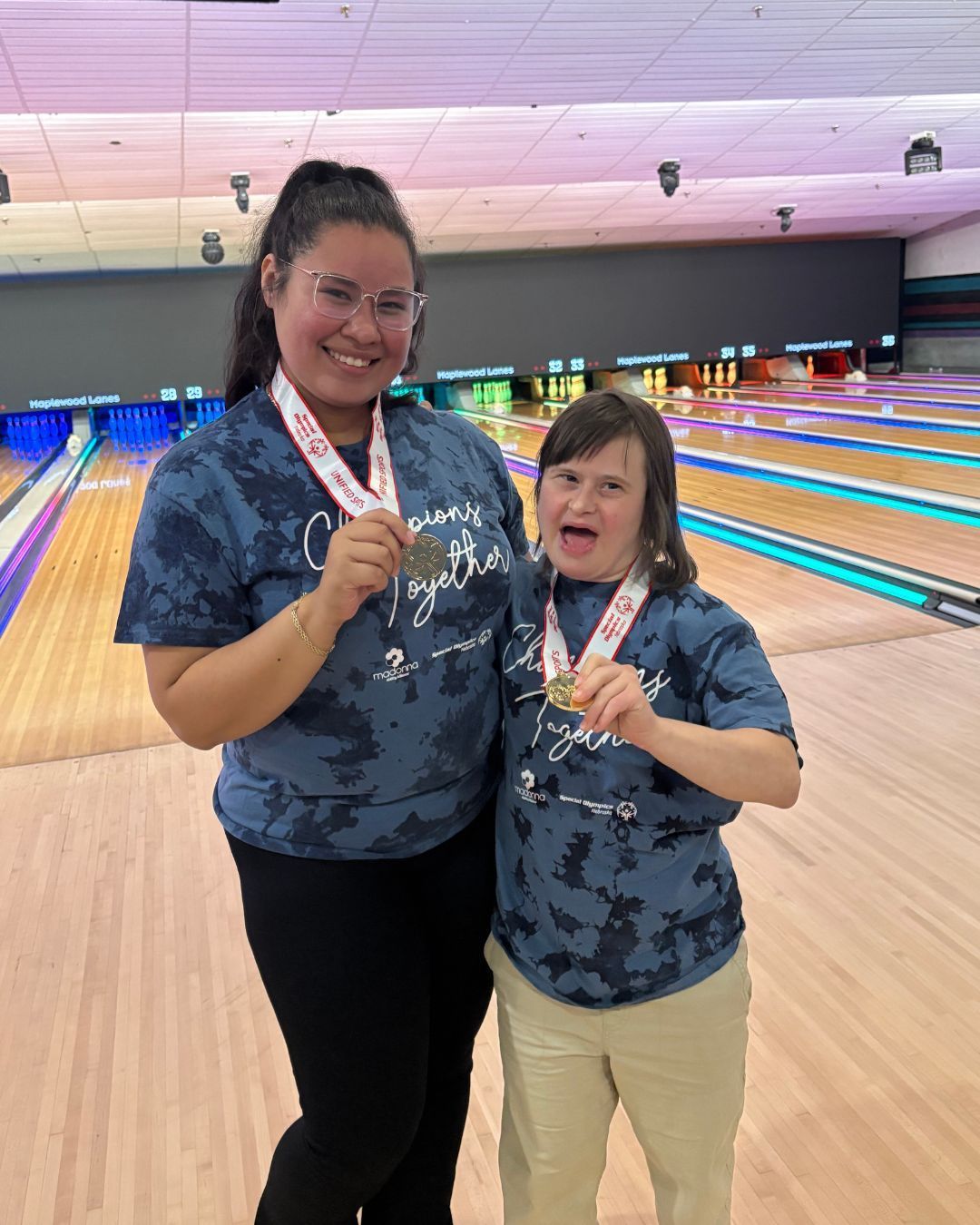 Two people smiling, holding medals, in a bowling alley. They wear matching blue tie-dye shirts.