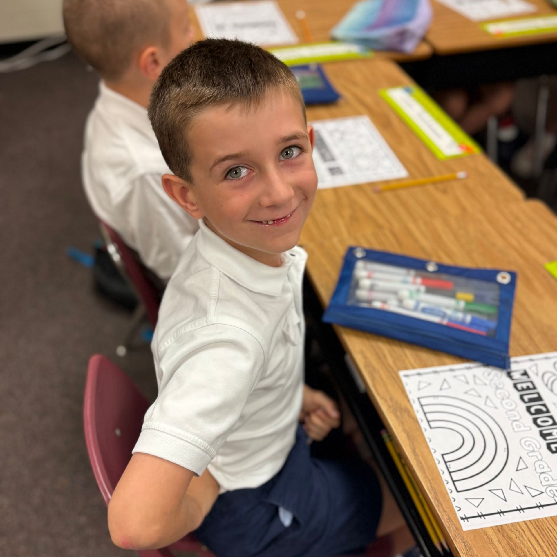 Boy in a white shirt and navy shorts smiles at the camera at his desk in class.
