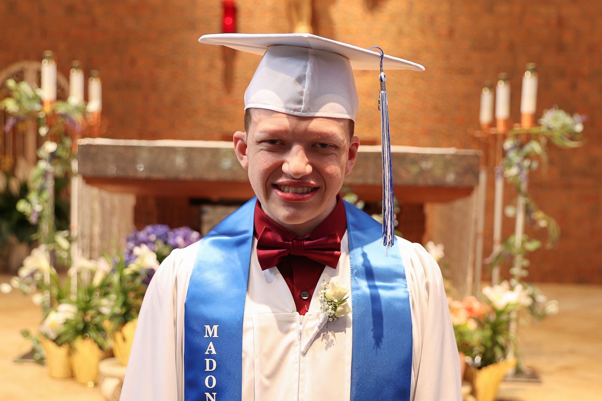 Man in graduation attire smiles, standing in a church. He wears a white cap and gown, blue sash, and maroon bow tie.