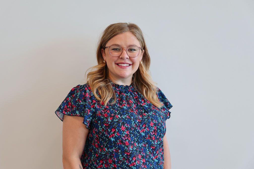 Woman in glasses and floral print dress smiles, standing in front of a neutral wall.