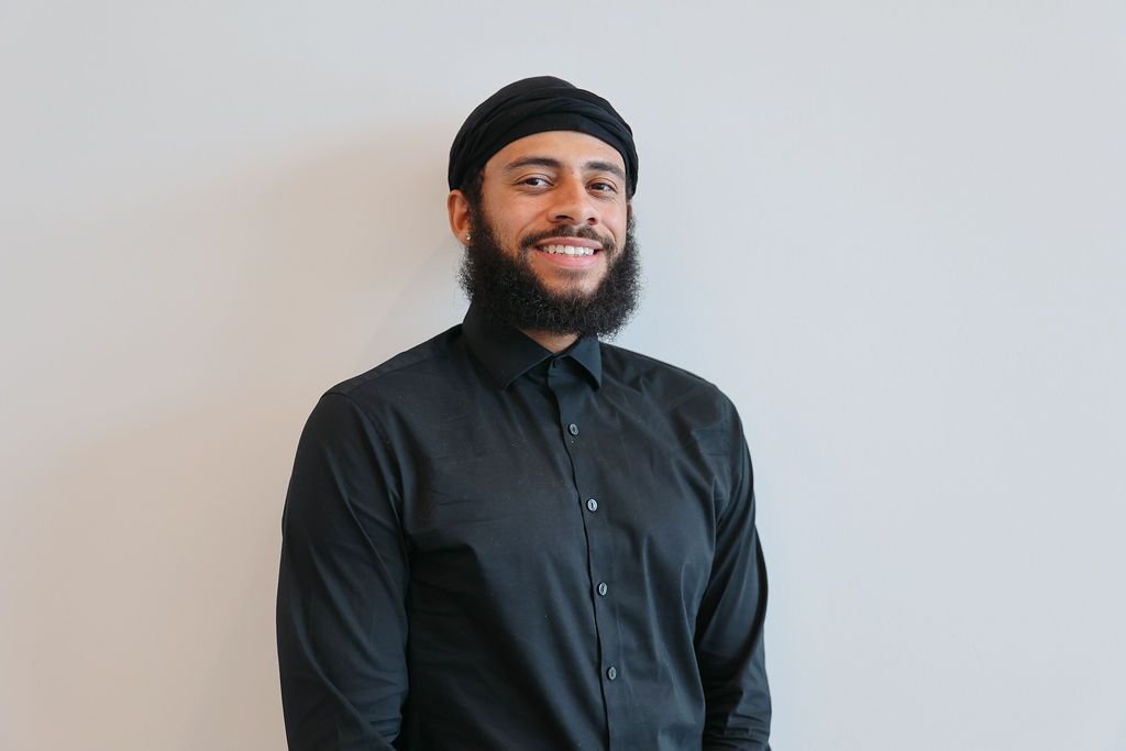 Man in black shirt and hat smiles against a white wall.