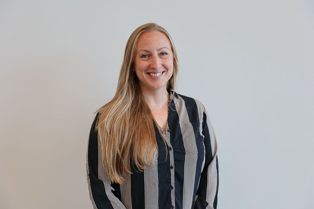 Woman with long blonde hair, smiling, wearing a striped shirt, standing in front of a white wall.