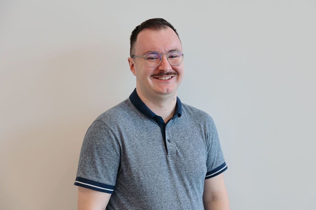 Man with glasses, mustache, and gray polo smiling in front of a neutral wall.