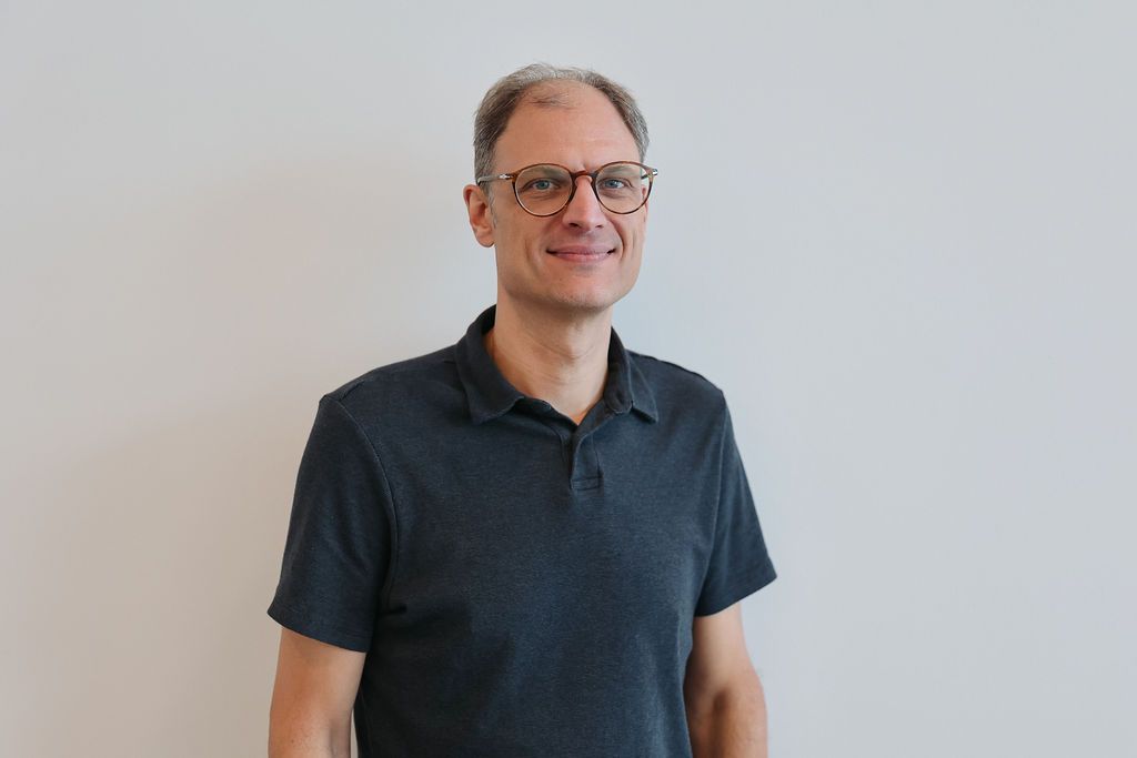 Man with glasses, gray hair, in dark polo shirt, smiling, standing against a white wall.