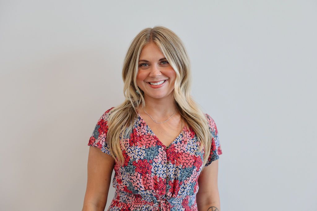 Woman smiling, wearing a floral dress, standing in front of a white wall.