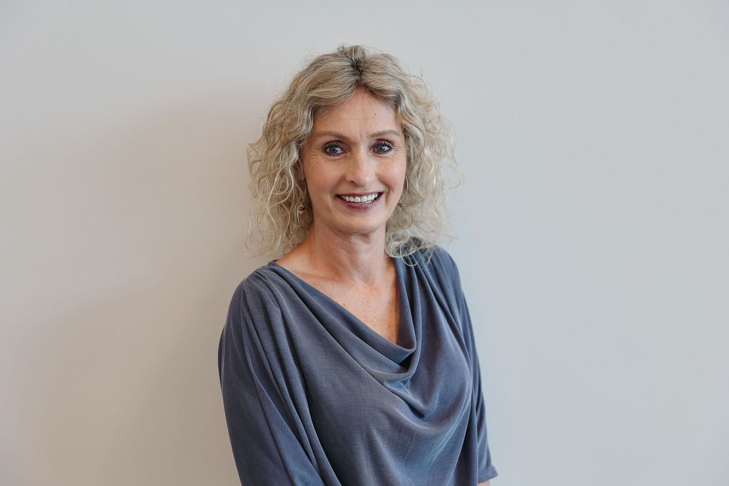 Woman with curly blonde hair smiles, wearing a blue-gray cowl neck top, against a white wall.