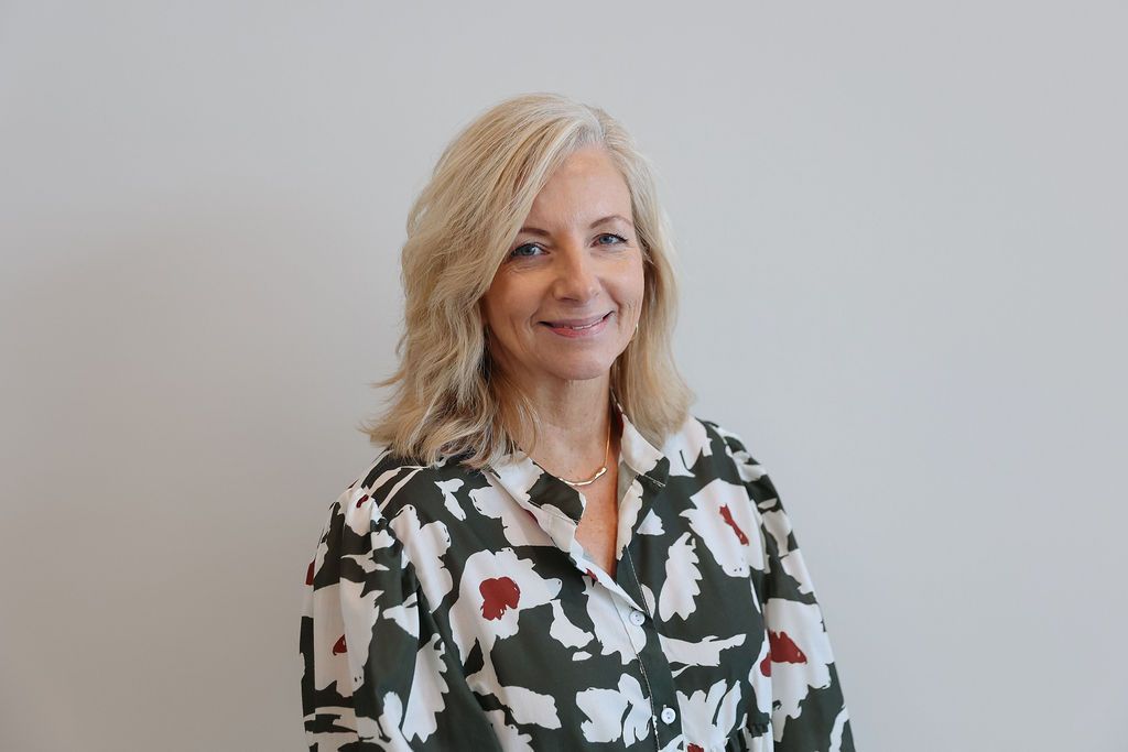 Woman with blonde hair wearing a patterned shirt, smiling. Standing in front of a white wall.