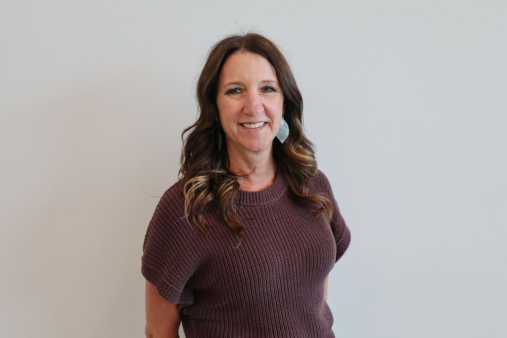 Woman with brown hair smiles, wearing a purple sweater, standing against a white wall.