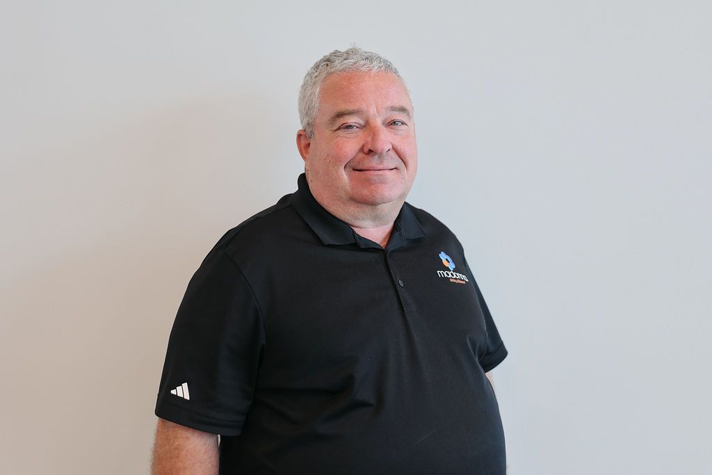 Man with gray hair wearing a black polo shirt, smiling, standing in front of a white wall.