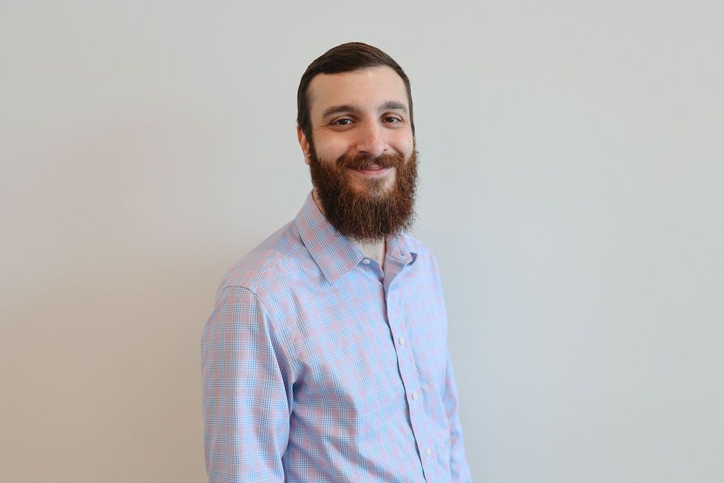 Man with a full beard smiling, wearing a blue plaid button-down shirt, against a plain background.
