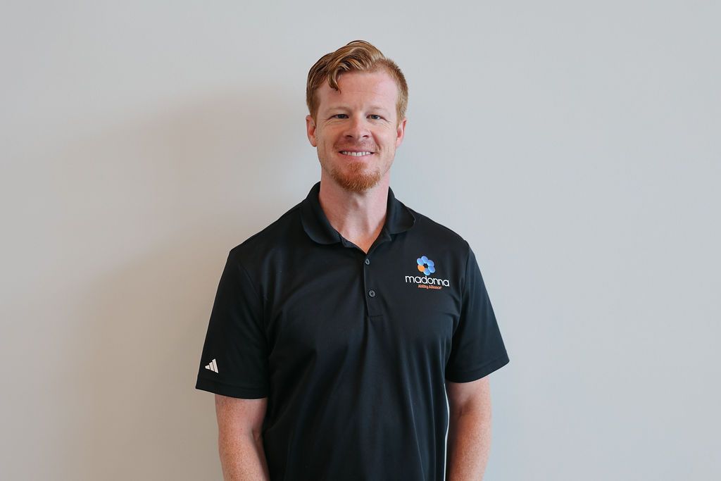 Man with red hair, wearing a black polo shirt, smiling, standing against a white wall.