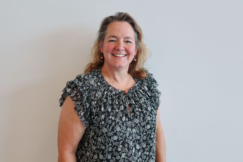 Woman with shoulder-length, light brown hair, smiles. She wears a black and white floral top against a white wall.