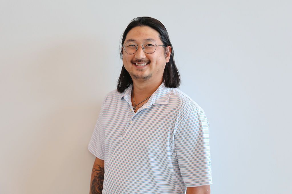 Man with long dark hair, wearing glasses and a striped shirt, smiling against a white wall.