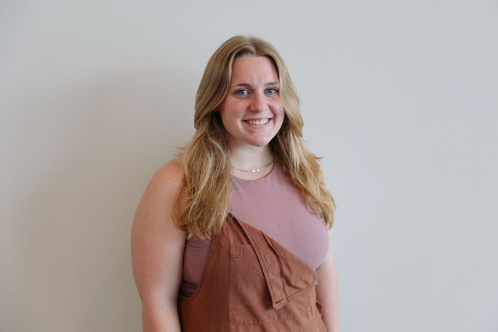 Woman with long blonde hair smiles, wearing a pink top and brown overalls. Against a white wall.