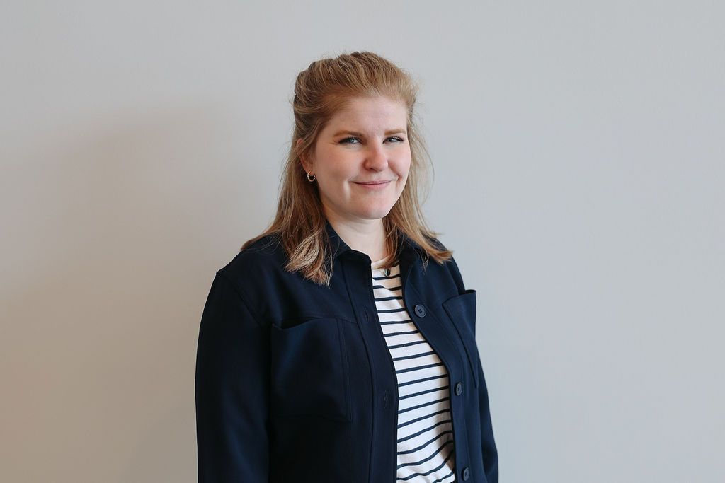 Woman with blonde hair wearing a striped shirt and dark blue jacket smiles in front of a gray wall.