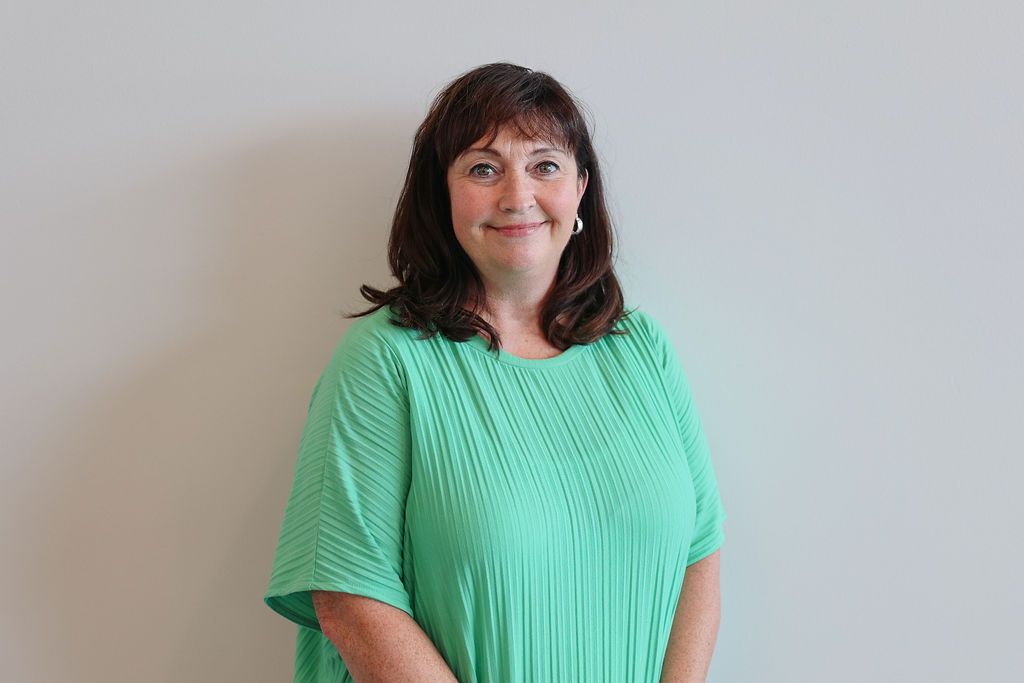Woman in green pleated top smiles at the camera, standing against a plain white wall.