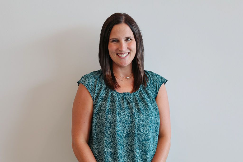 Woman in a teal patterned top smiles, standing against a plain white wall.