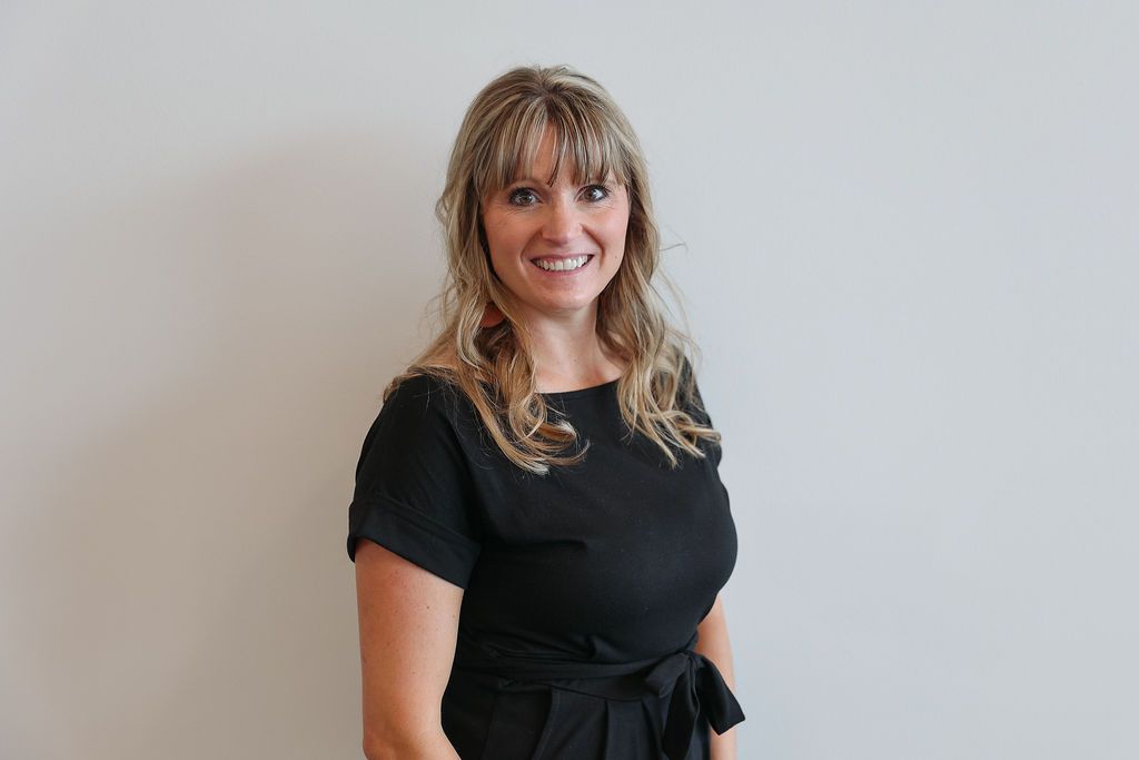 Woman in black top smiles against a white wall.