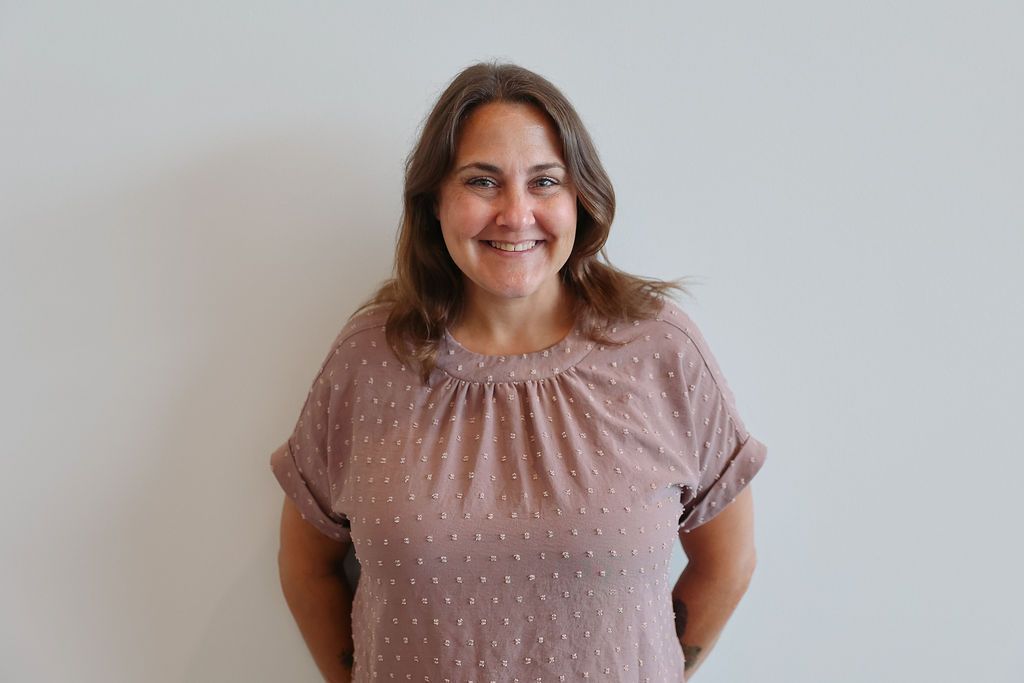 Woman smiling, standing in front of a white wall, wearing a brown textured blouse, arms behind back.