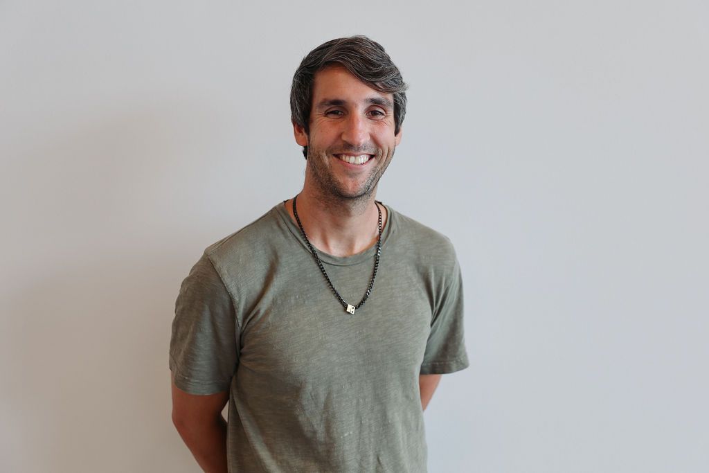 Man in a green shirt smiles at the camera, hands behind back, in front of a white wall.